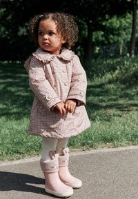 Manteau imperméable à carreaux roses avec col à volants, associé à des leggings crème et des bottes de pluie roses ornées d'accents en forme d'oreilles de lapin.