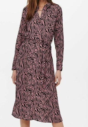 Kobieta w różowo-czarnej sukience w paski zebra, z długim rękawem, sięgającej do kolan, zapinanej na guziki, z paskiem wiązanym w talii.