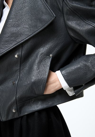 Persoon die een zwarte leren jas met textuur draagt en zilveren drukknopen, met de hand in de zijzak en zichtbaar witte overhemdmanchetten.