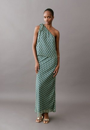 Femme portant un haut asymétrique vert et blanc à motifs et une jupe longue assortie, debout devant un fond gris.
