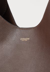 Τσάντα χειρός από σκούρο καφέ δέρμα Coach New York που δείχνει λεπτή ραφή και χρυσλό λογότυπο σε λείο υφασμένο φόντο.