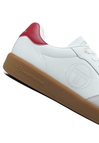 Bílá teniska s červeným detailem na patě, texturovanou gumovou podrážkou a jemným logem na boku. Šněrovací design.