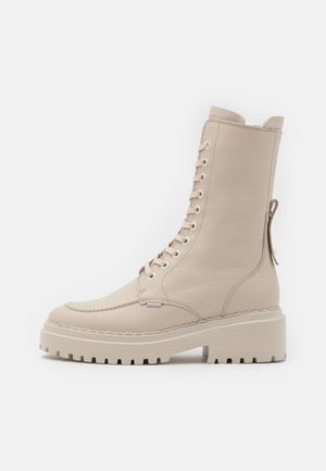 Beigen värinen nahkainen nyörillinen taistelukenkä, jossa on paksu uritetulla kuvioinnit pohja, pyöreä kärki, vetokahva kantapäässä ja tikattu yksityiskohta, esitetty sivulta valkoisella taustalla.