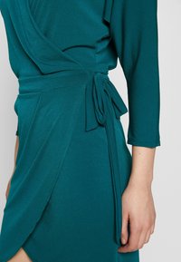 Teal omslagsklänning med långa ärmar, med en sidoknut, draperad tyg och slät yta. Designad för en figurnära silhuett.