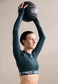 Zielona sportowa bluzka z długim rękawem o skróconym kroju, z białą taśmą "NIKE PRO". Model trzyma czarną piłkę lekarską nad głową.