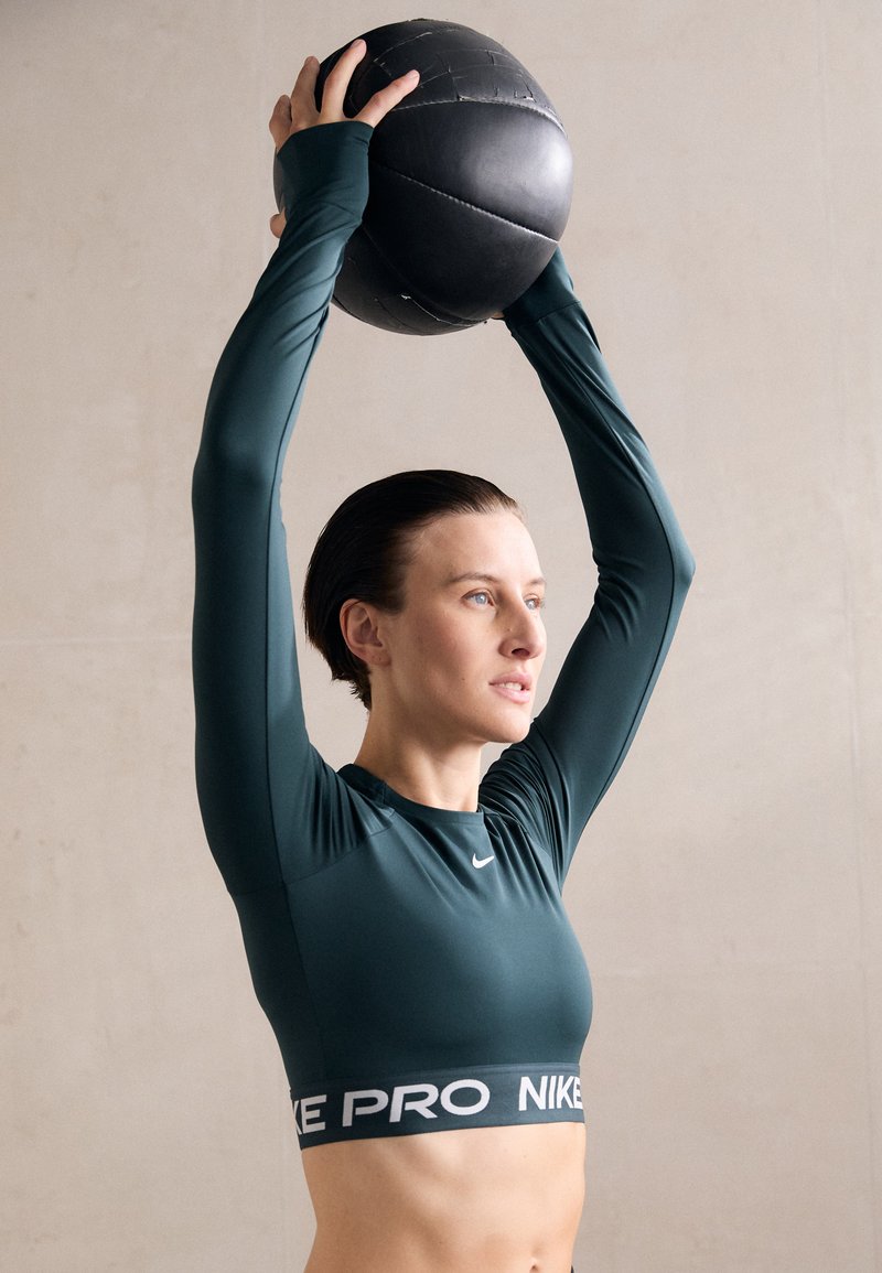 Zielona sportowa bluzka z długim rękawem o skróconym kroju, z białą taśmą "NIKE PRO". Model trzyma czarną piłkę lekarską nad głową.