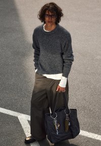 Suéter de punto gris, camisa blanca debajo, pantalones anchos de oliva, bolsa tote negra con detalles de metal y llaveros colgantes. Atuendo casual y texturizado.