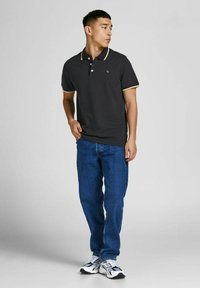 Dark gray polo shirt with a yellow collar accent, three buttons, paired with blue jeans and white sneakers. Model stands with one hand on hip.