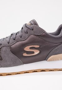 Baskets Skechers grises avec empiècements en daim et mesh, logo "S" beige, semelle texturée et fermeture à lacets sur fond blanc.