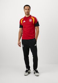 Homem em pé, vestindo uma camisola de futebol da Alemanha em vermelho e preto e calças de treino pretas da Adidas com ténis brancos, contra um fundo branco liso.