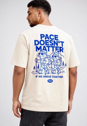 Man wearing a beige T-shirt with blue text "PACE DOESN'T MATTER IF WE ARRIVE TOGETHER" and hiking cartoon animals on the back.