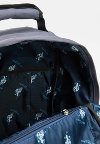 Vnitřek šedého batohu ukazující tmavě modrou látkovou podšívku s opakujícím se světle modrým logem U.S. Polo Assn. a přihrádkou na zip.