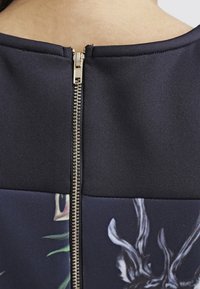 Lahti sininen paita, jossa on takana kultainen metallivetoketju, ja tekstuurinen kangas, joka kontrastiin painetun alaosan, jossa on hirviä kuva.