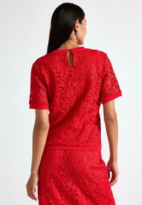 Kvinde med langt mørkt hår iført en kortærmet rød blonde-top og en matchende nederdel, vendende væk mod en hvid baggrund.