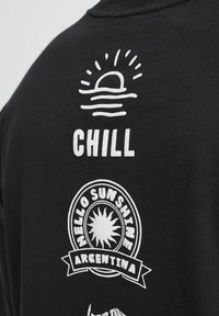 Černé bavlněné tričko s bílým sítotiskovým potiskem. Design zahrnuje slunce a vlny, s textem: "CHILL" a "AHOJ SLUNÍČKO ARGENTINA."