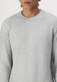Pull en tricot côtelé gris avec un col rond et des manches longues, présentant un motif texturé et une silhouette ajustée.