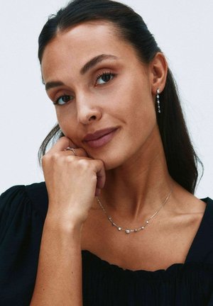 Femme aux cheveux foncés porte des boucles d'oreilles pendantes, un collier délicat avec de petites pierres, un haut noir, posant le menton sur la main, fond neutre.