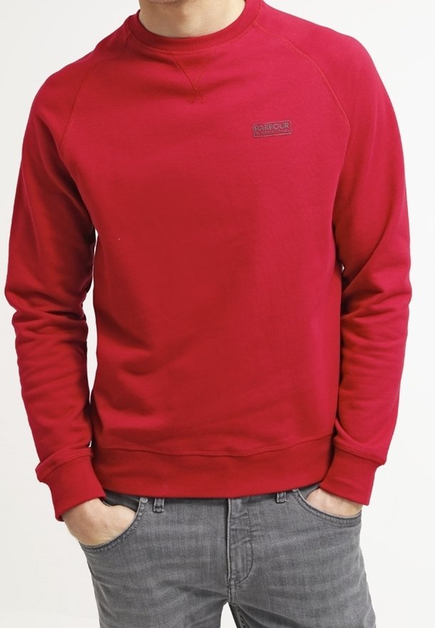 Homme portant un sweat-shirt rouge uni à manches longues avec un petit logo Barbour sur la poitrine et un jean gris, les mains dans les poches.