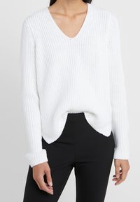 Biały dziergany sweter z dekoltem w serek, o fakturze prążkowanej, z długimi rękawami i zaokrąglonym dołem, zestawiony z czarnymi spodniami.