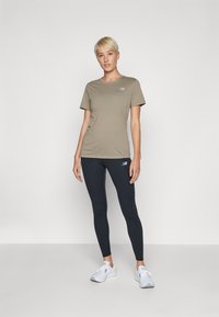 T-shirt kaki à manches courtes avec un petit logo, associé à des leggings noirs, complété par des chaussures de sport blanches. Design simple et ajusté.