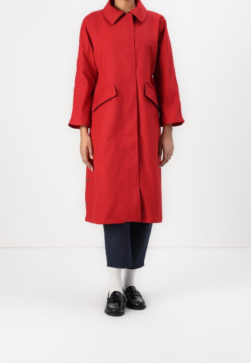 Femme portant un manteau rouge mi-long avec des poches à rabat, un pantalon noir, des chaussettes blanches et des mocassins noirs, debout devant un fond uni.