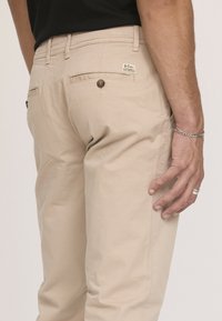 Pantalons en coton beige au design ajusté, avec des poches arrière, une petite étiquette et un détail de bouton. Texture lisse et style décontracté.