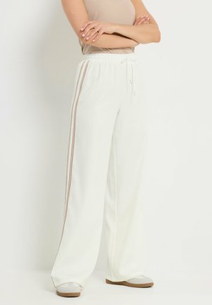 Osoba ubrana w beżowy top bez rękawów oraz białe szerokie spodnie z brązowymi paskami po bokach i białe tenisówki, stojąca z założonymi rękami.