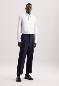 Camicia bianca a maniche lunghe con una tasca abbinata a pantaloni blu scuro con pieghe e scarpe eleganti nere. Tessuto liscio e vestibilità sartoriale.