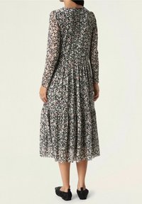 Vestido negro con flores, de mangas largas, tejido transparente, falda plisada y un colorido patrón floral con acentos en rosa y blanco. Lleva zapatos negros.