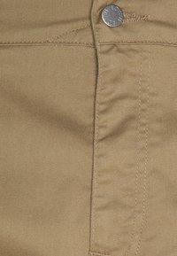 Bežová bavlněná látka s diagonálním žebrováním, viditelným stehem a kovovým knoflíkem na pásku.