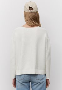 Weißer, gerippter Pullover mit lockerem Sitz, rundem Ausschnitt und abfallenden Schulternähten, kombiniert mit hellblauen Jeans und einer beigen Kappe.