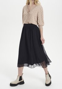 Beige knitted cardigan with short sleeves, paired with a black, flowing skirt and beige ankle boots with black soles and side elastic.