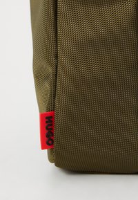 Olivenfarget teksturert bag med et rødt merke. Stoffet har et mønster av fine linjer, og bagen har en strukturert form.