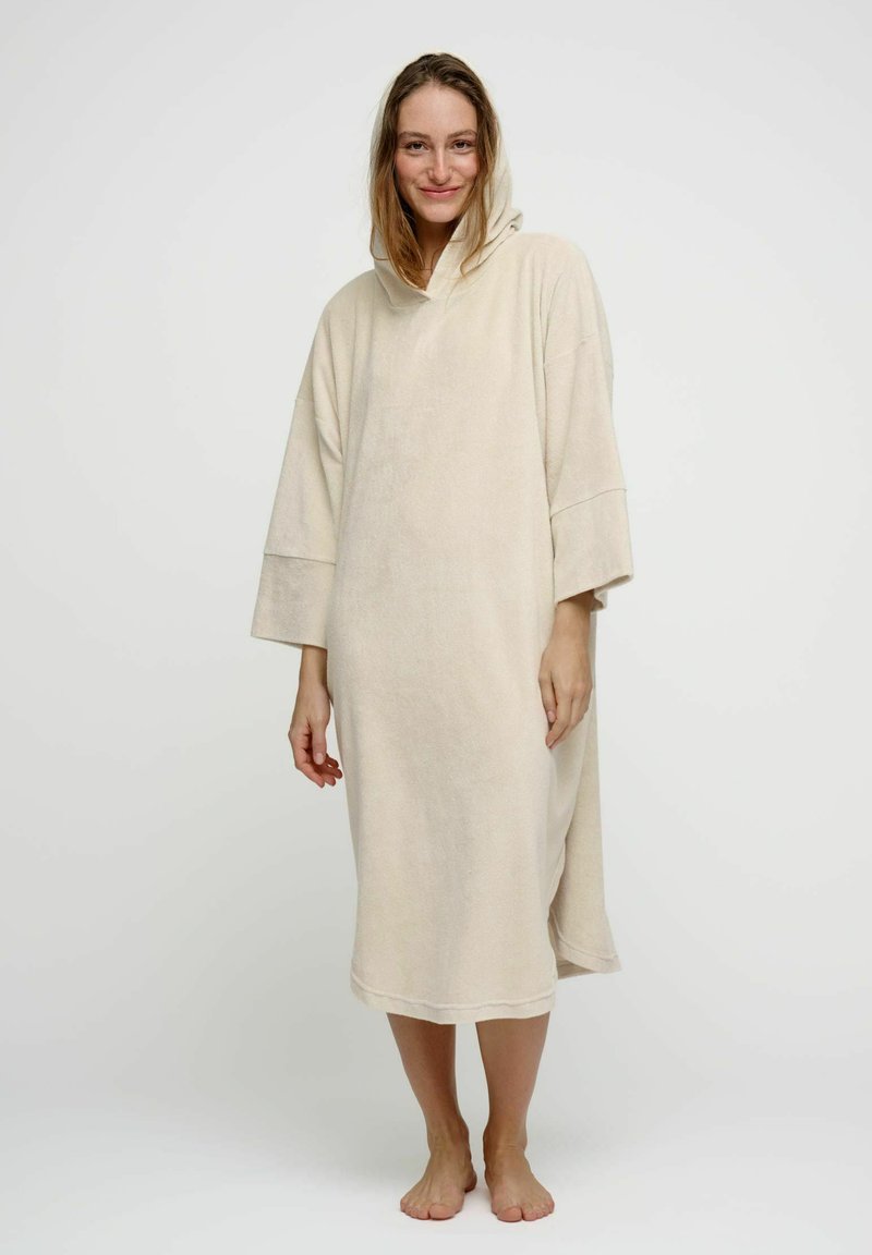 Femme portant une robe à capuche ample beige, debout pieds nus devant un fond blanc uni, souriant doucement à la caméra.