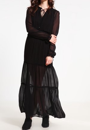 Frau trägt ein langärmliges, schwarzes, durchsichtiges Maxikleid mit Stiefeletten und steht vor einem schlichten weißen Hintergrund.