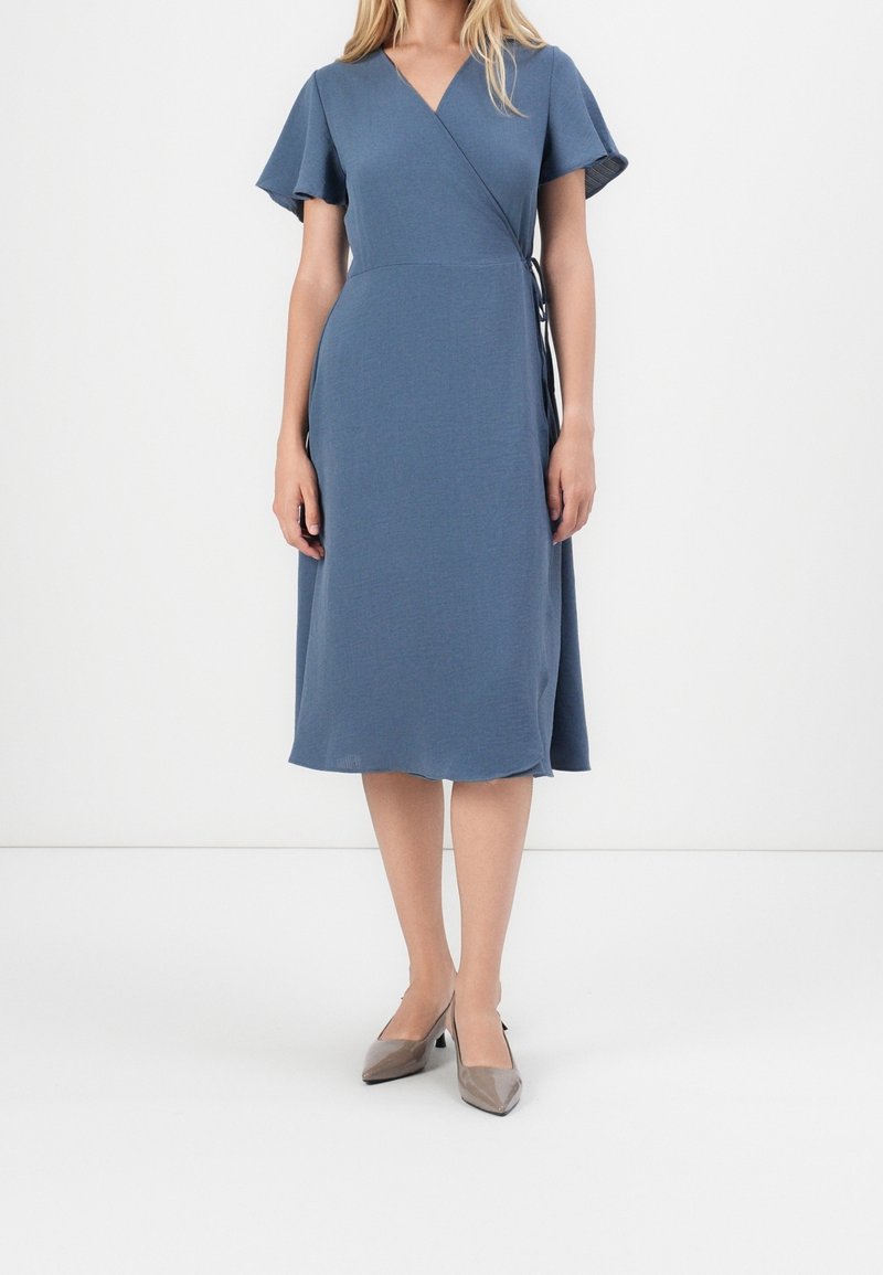 Femme vêtue d'une robe portefeuille bleue avec des manches courtes volantées et des escarpins gris à bouts pointus, debout sur un fond blanc uni.
