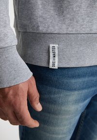 Harmaa collegepaita, jossa on ribbireunat hihansuissa ja helmassa, ja jossa on kangasilmätieto, jossa lukee "DREI MASTER." Henkilön käsi lepää sinisten farkkujen päällä.
