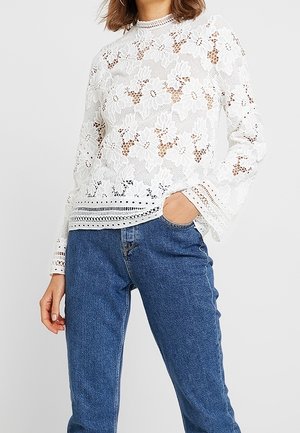 Woman wearing a white floral lace long-sleeve blouse and blue high-waisted jeans, with one hand in her pocket.