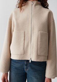Giacca a zip in lana beige con due grandi tasche frontali, indossata con jeans blu, mostrata dalla metà del torso al collo.