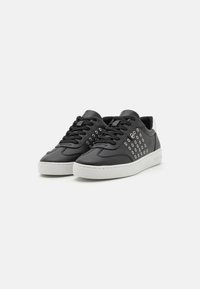 Sneaker in pelle nera con suola bianca, caratterizzate da occhielli metallici decorativi sui lati e lacci neri. Texture liscia e forma arrotondata.