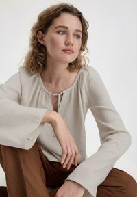 Blusa beige chiaro con scollatura rotonda, dettaglio a chiave e maniche lunghe a sbuffo, abbinata a pantaloni marroni. Tessuto morbido e strutturato.