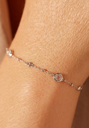 Silver bracelet with linked chain, featuring clear gemstones set in round bezels, resting on a textured skin surface.