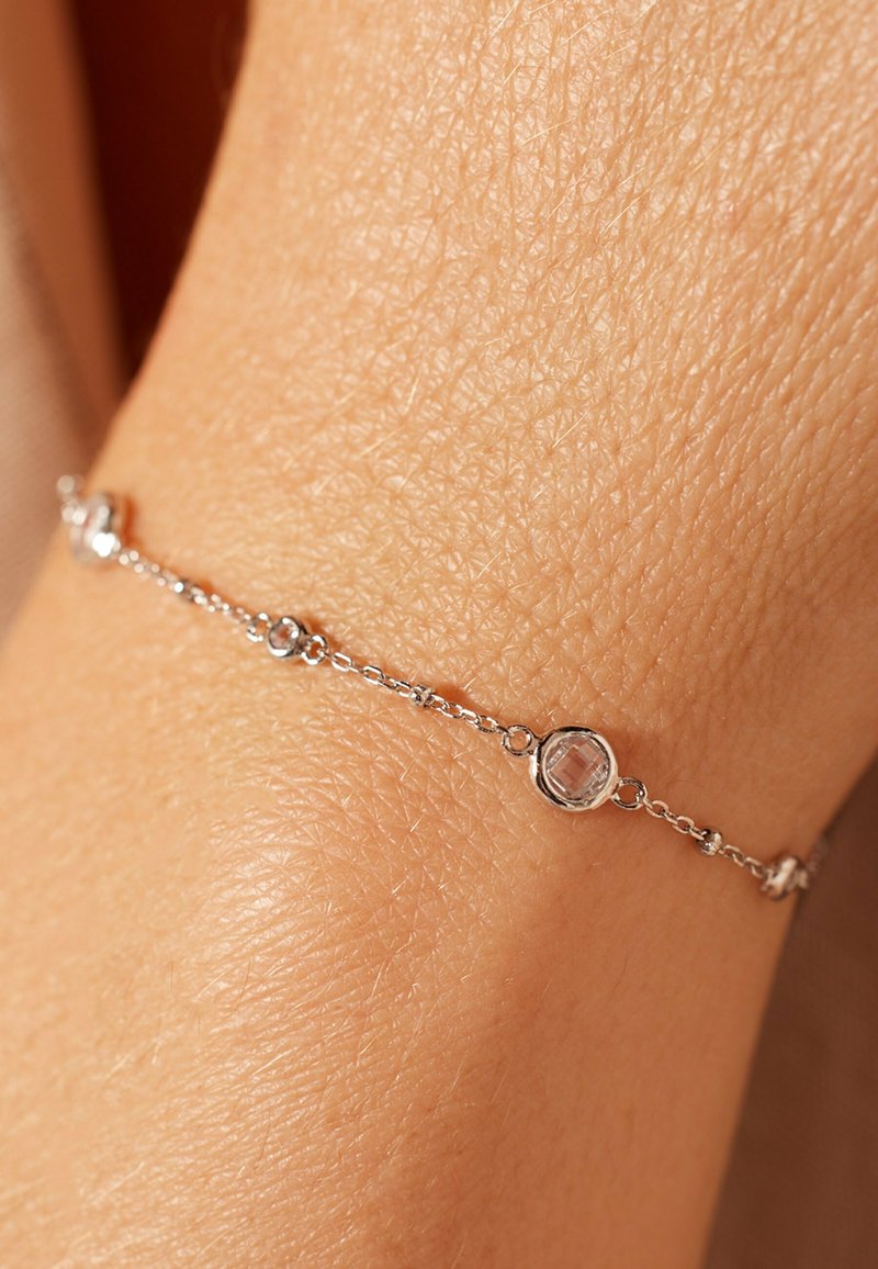 Zilveren armband met een schakelkettinkje, voorzien van heldere edelstenen in ronde zettingen, rustend op een textuur van de huid.