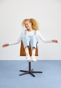 Young woman with curly hair smiles with eyes closed while balancing on a wooden swivel chair in a minimalist room.