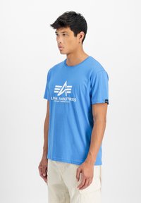 Modrá krátkostranná trička vyrobená z měkké tkaniny, s bílým logem se šikmými vzory. Kulatý výstřih; štítek na levém rukávu.