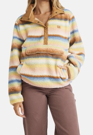 Femme portant un pull polaire à rayures multicolores avec un col à boutons partiels et une poche kangourou, assorti à un pantalon marron.