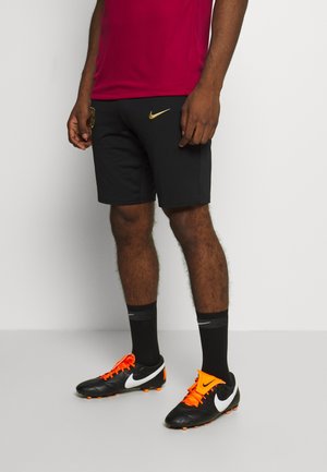 Mann trägt schwarze Shorts mit goldenem Nike-Logo, schwarze Socken und schwarze Fußballschuhe mit weißem Swoosh und leuchtend orangefarbenen Schnürsenkeln, steht auf grauem Boden.