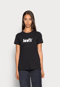 Camiseta de algodón negra con un logo blanco de Levi's en el frente, cuello redondo, mangas cortas y corte casual. Modelo mostrado con pantalones de cuadros.