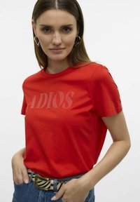 T-shirt en coton rouge à manches courtes, avec le mot "ADIOS" imprimé en une teinte plus claire sur le devant, associé à un jean en denim.