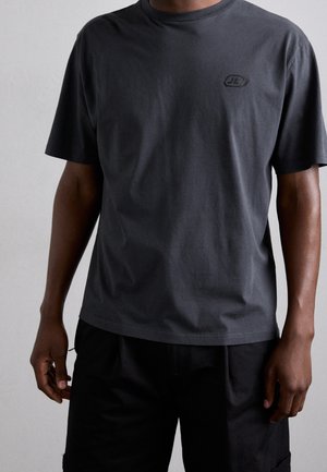 Uomo che indossa una semplice t-shirt a maniche corte grigio scuro con un piccolo logo discreto sul lato sinistro del petto e pantaloncini neri.